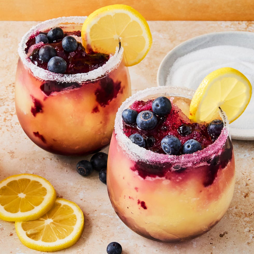 Frozen Blueberry Lemon Drop Slushies stemless glasses filled with a frozen vodka lemon cocktail with a blueberry syrup swirled in the glass. garnished with a sugared rim, lemon wheels, and fresh blueberries