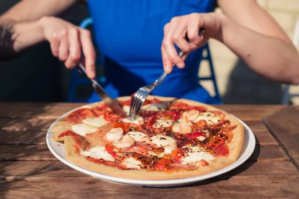 A young woman in a blue dress is sitting outside at a restaurant table and is cutting a pizza