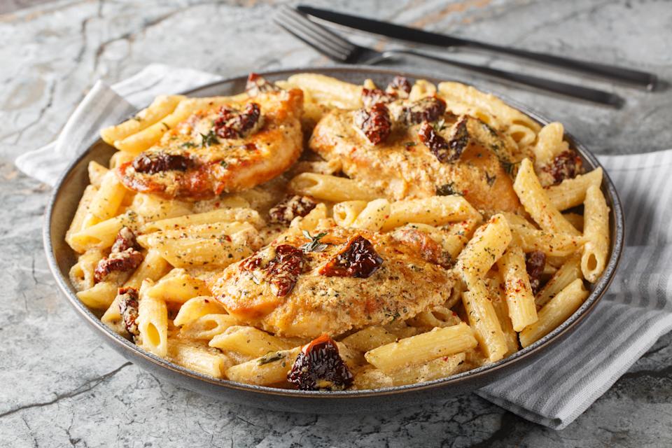 Pasta dish with grilled chicken, sun-dried tomatoes, and creamy sauce on a plate, placed on a marble surface with cutlery nearby