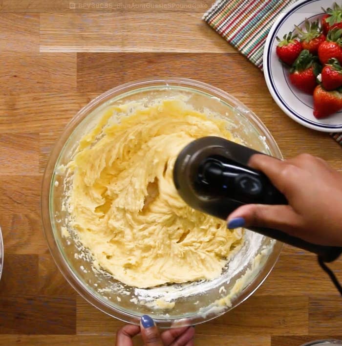 Person mixing cake batter in a glass bowl with a hand mixer, strawberries nearby on a plate