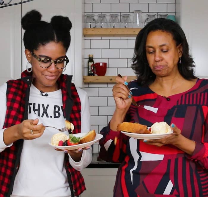 Two people enjoying cake with ice cream in a kitchen, wearing casual attire. Shelves with kitchen items are visible in the background