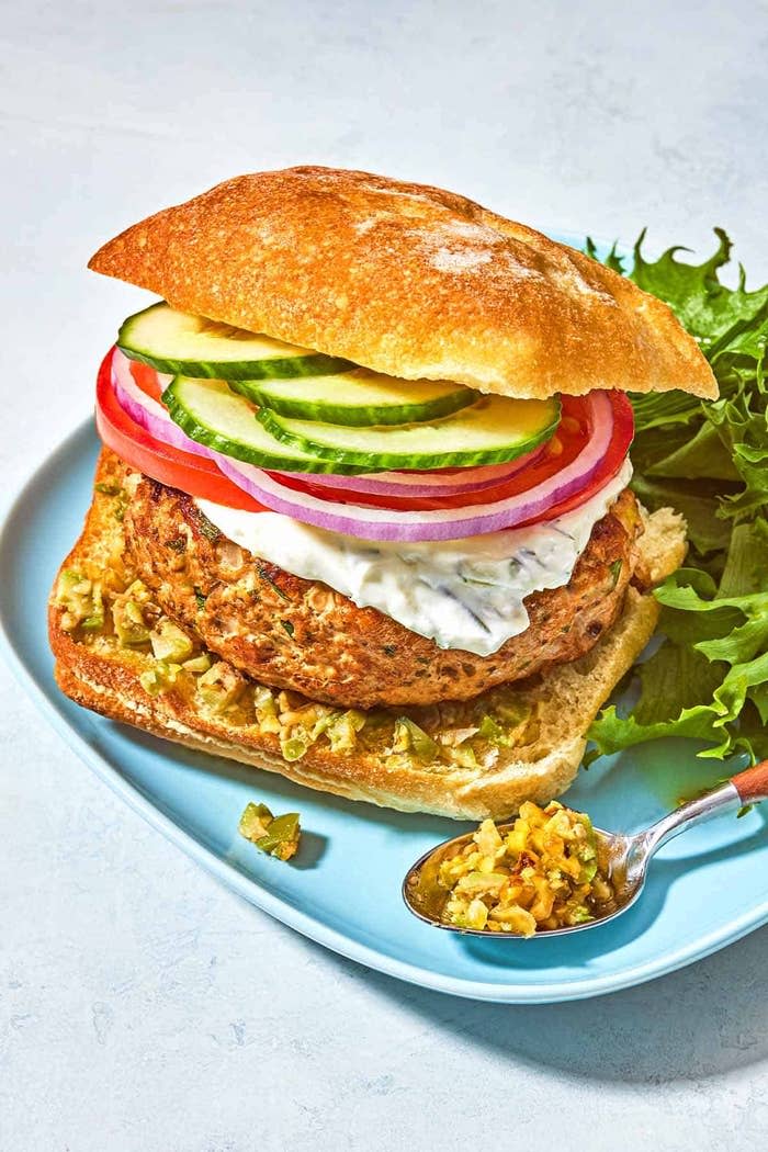 Gourmet burger with sliced cucumbers, tomatoes, onions, and tzatziki sauce on ciabatta bread, served with a leafy greens garnish on a light blue plate