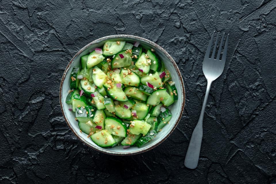 Bowl of cucumber salad with diced onions and seasoning, on a textured dark surface, next to a fork