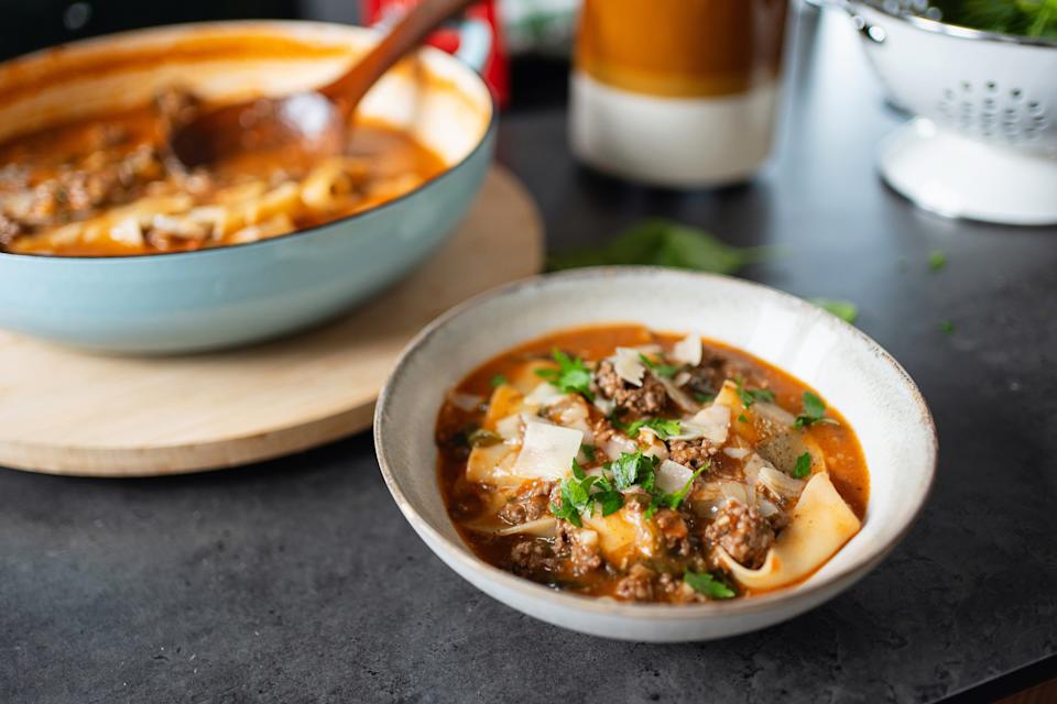 Bowl of lasagna soup with noodles, meat, and cheese, garnished with herbs. A pot of soup is in the background