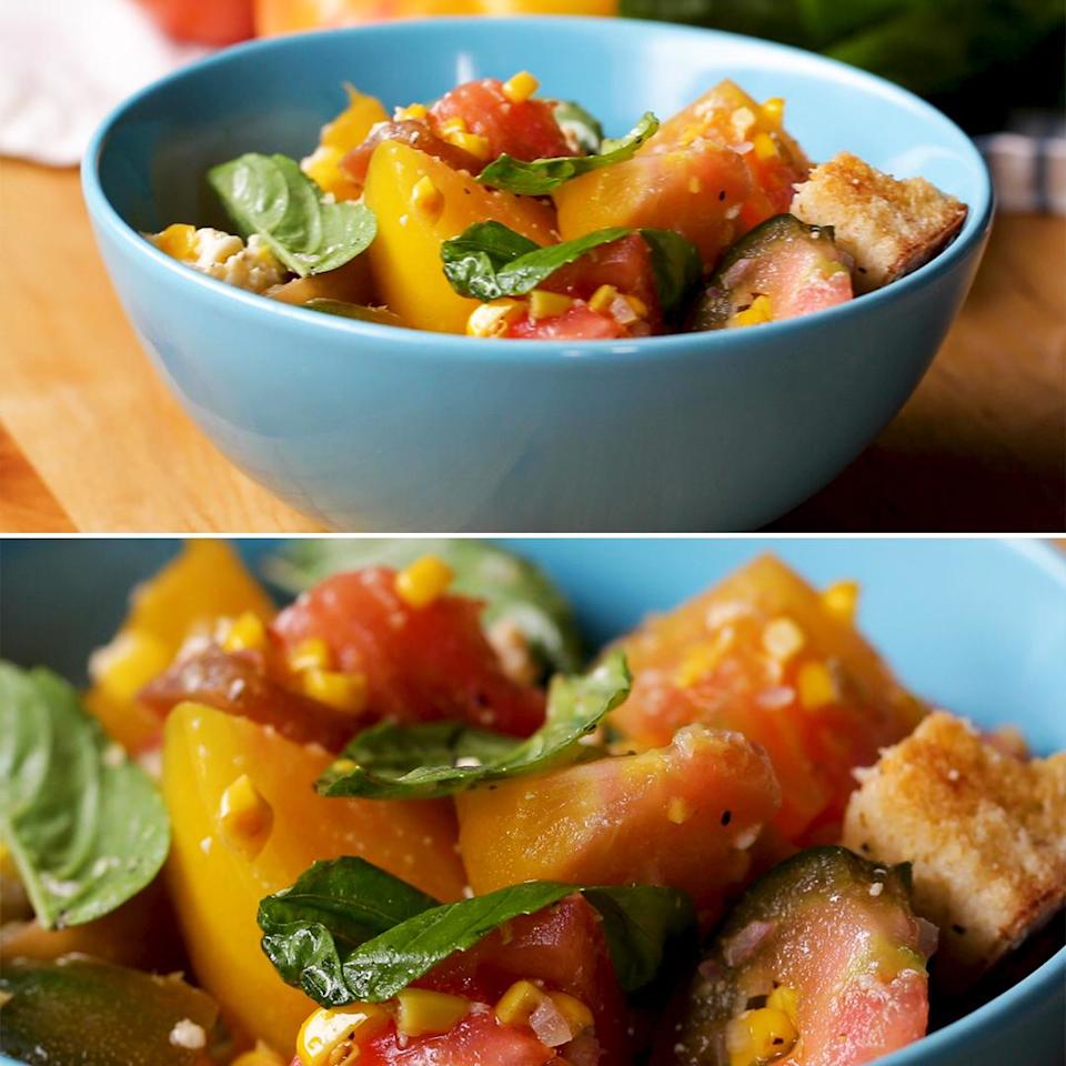A close-up of a colorful salad with tomatoes, basil, bread pieces, corn, and a touch of olive oil, served in a blue bowl