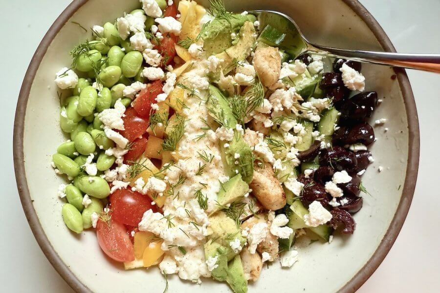 Just in Time for June: Two New Lazy Girl High-Protein Salads (Topped with Chips!) - 9 A bowl filled with edamame, tomatoes, yellow peppers, cucumber, olives, avocado, chicken, feta cheese, and dill offers a high-protein delight. Topped with cottage cheese for added richness and fiber, it's served invitingly with a spoon.
