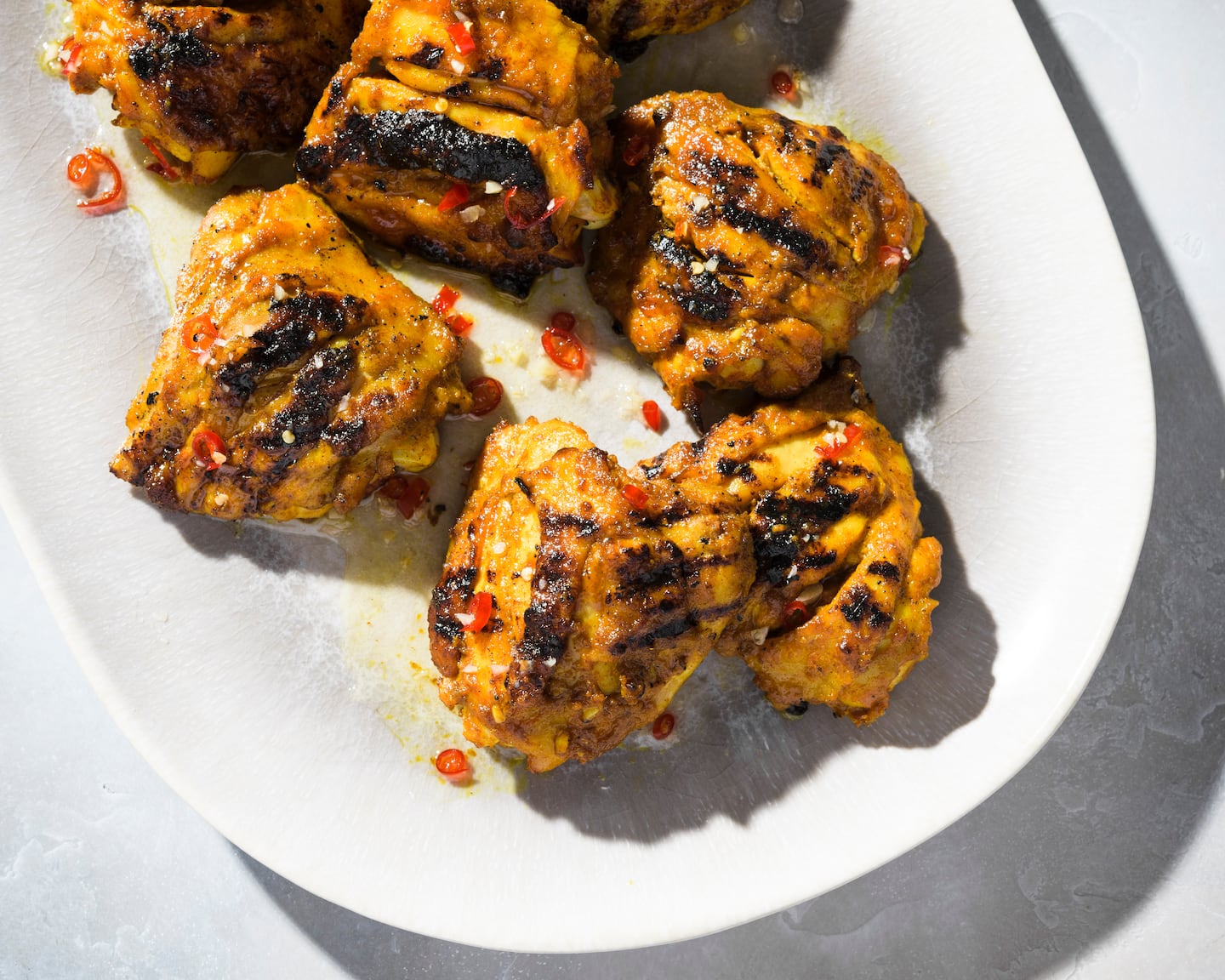 A white serving platter with about a half-dozen grilled chicken thighs topped with some red peppers.