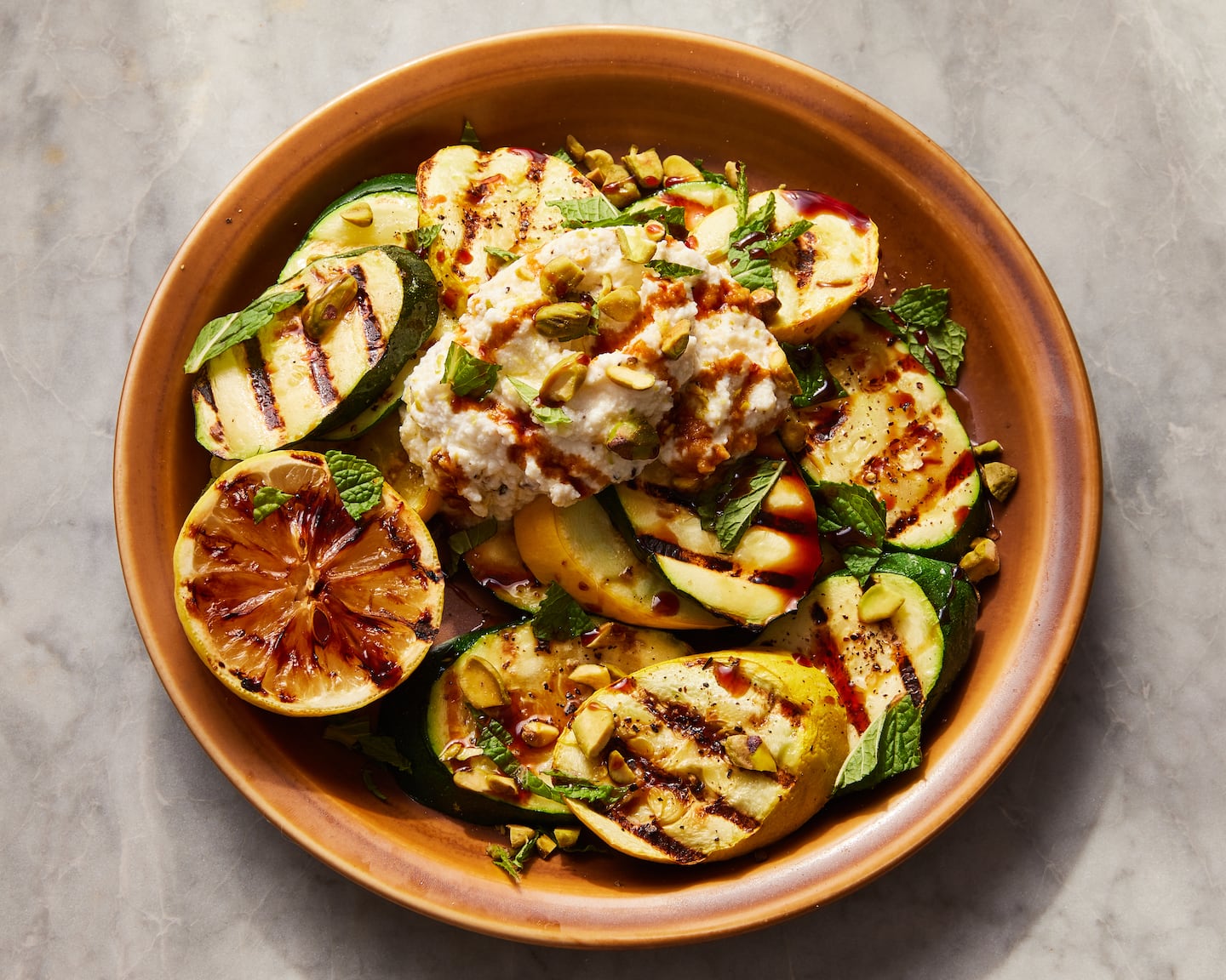 A bowl of grilled zucchini garnished with green herbs and a grilled half lemon.
