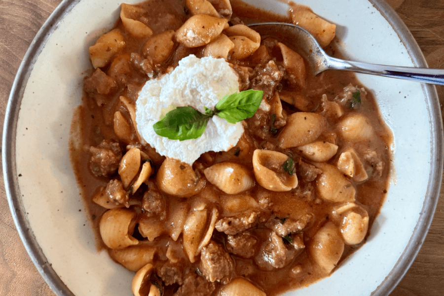 Just in Time for June: Two New Lazy Girl High-Protein Salads (Topped with Chips!) - 7 A plate of pasta shells in a rich tomato sauce with ground meat resembles a deconstructed lasagna, topped with creamy ricotta cheese and a fresh basil leaf. A spoon is on the plate.