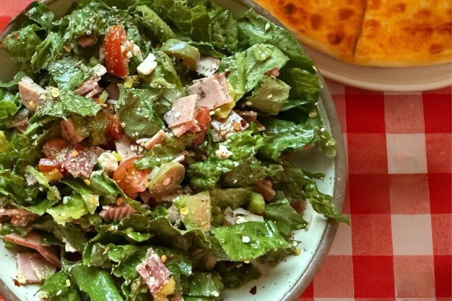Just in Time for June: Two New Lazy Girl High-Protein Salads (Topped with Chips!) - 4 A delicious salad with leafy greens, chopped tomatoes, sliced meat, and cheese sits elegantly on a plate. Hinting at rustic charm, part of a round bread rests nearby on the classic red and white checkered tablecloth—your perfect recipe for a refreshing meal.
