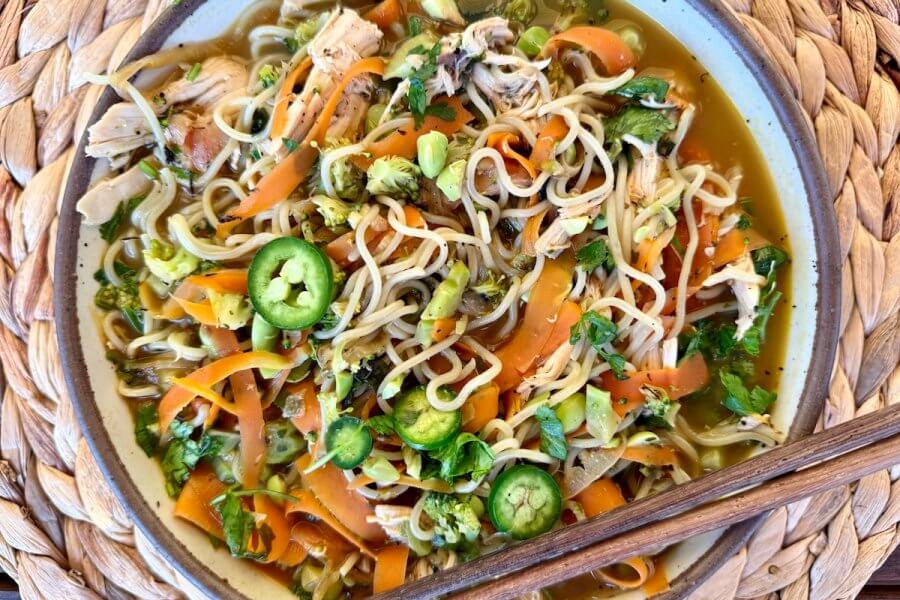 Just in Time for June: Two New Lazy Girl High-Protein Salads (Topped with Chips!) - 3 A high protein noodle bowl featuring shredded chicken, sliced carrots, cilantro, and jalapeño slices, served with chopsticks on a woven placemat—perfect for busy day meals.