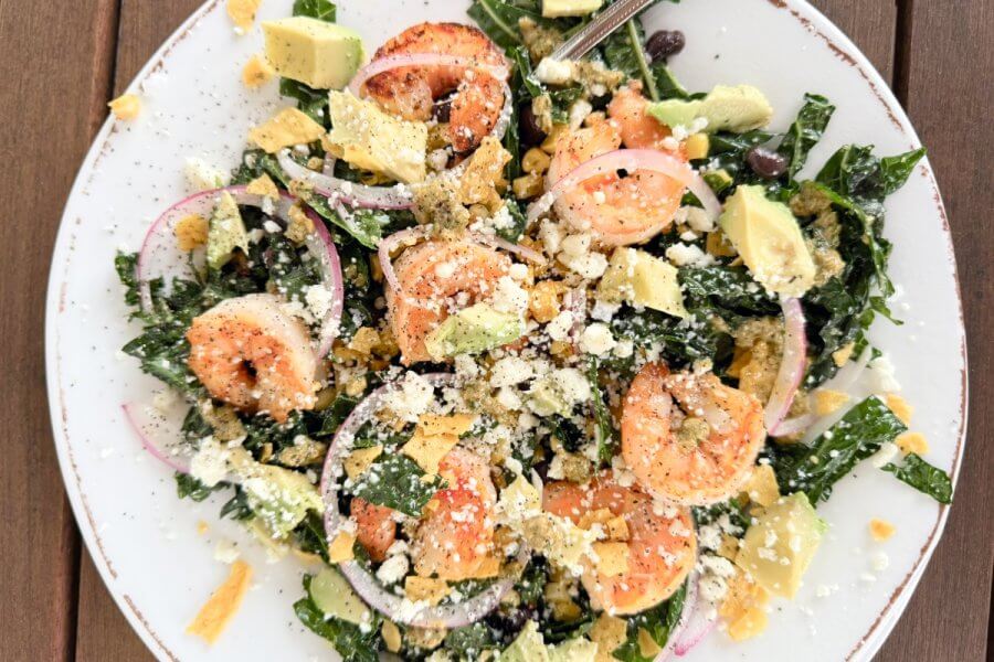 Just in Time for June: Two New Lazy Girl High-Protein Salads (Topped with Chips!) - 1 A plate of Shrimp Kale Salad with avocado, black beans, red onions, crumbled cheese, tortilla chips, and a zesty Lime Pepita Dressing sits on a wooden table.