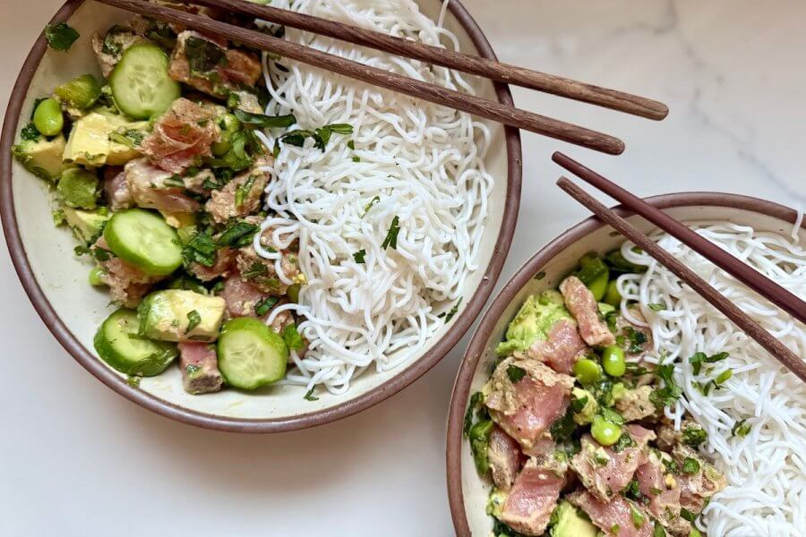 Just in Time for June: Two New Lazy Girl High-Protein Salads (Topped with Chips!) - 6 Two bowls containing rice noodles and a high-protein mix of raw fish, avocado, cucumber, and greens sit elegantly on a marble surface, with chopsticks resting on the edges.