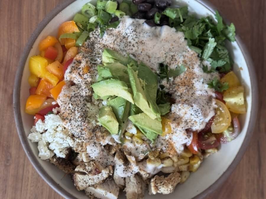 Just in Time for June: Two New Lazy Girl High-Protein Salads (Topped with Chips!) - 5 A bowl brimming with diced vegetables, black beans, grilled chicken, chopped greens, and avocado slices drizzled with creamy dressing—a perfect choice for those seeking high protein recipes.