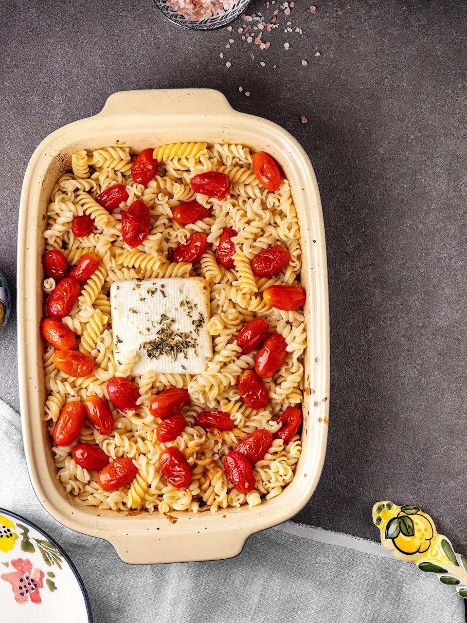 Baked pasta dish in a beige casserole with cherry tomatoes and a block of cheese topped with herbs