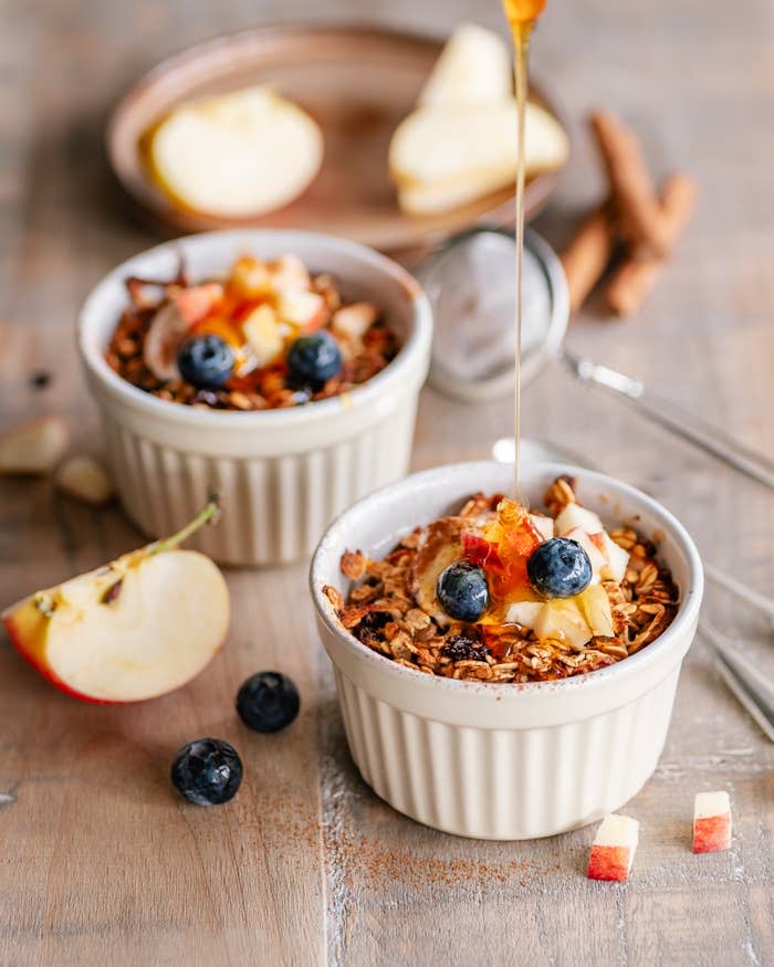 Two ramekins of granola topped with blueberries, sliced apples, and honey drizzle, set on a wooden table with fresh fruit slices and cinnamon sticks nearby