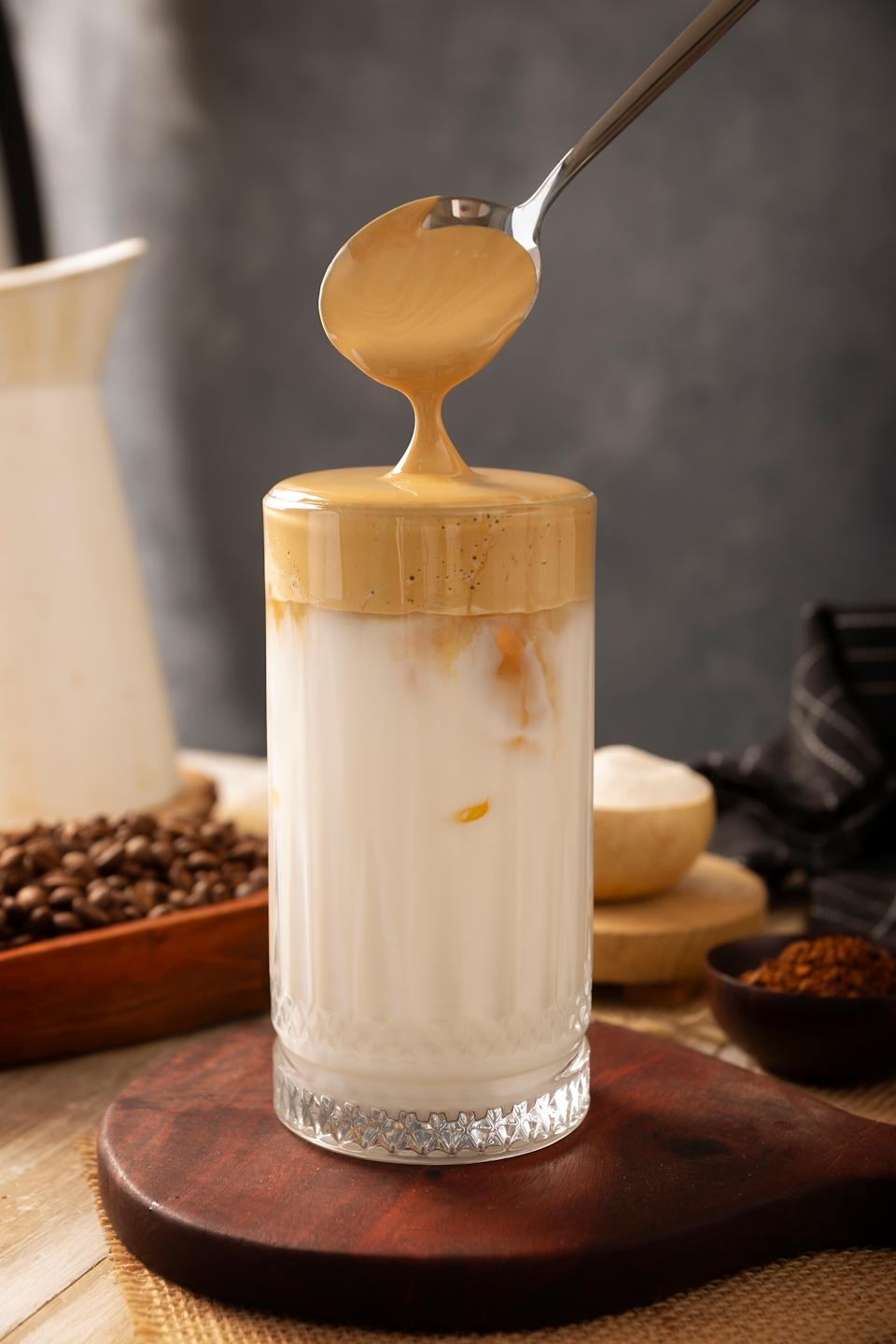 A spoon drizzles whipped coffee over a glass of iced milk, surrounded by coffee beans and powder in a cozy setting