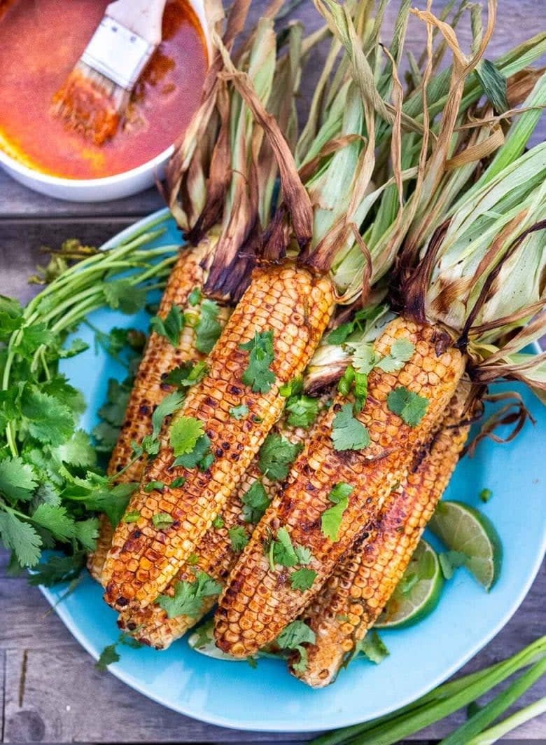 Grilled corn on a plate garnished with cilantro and lime, accompanied by a bowl of spicy sauce with a basting brush