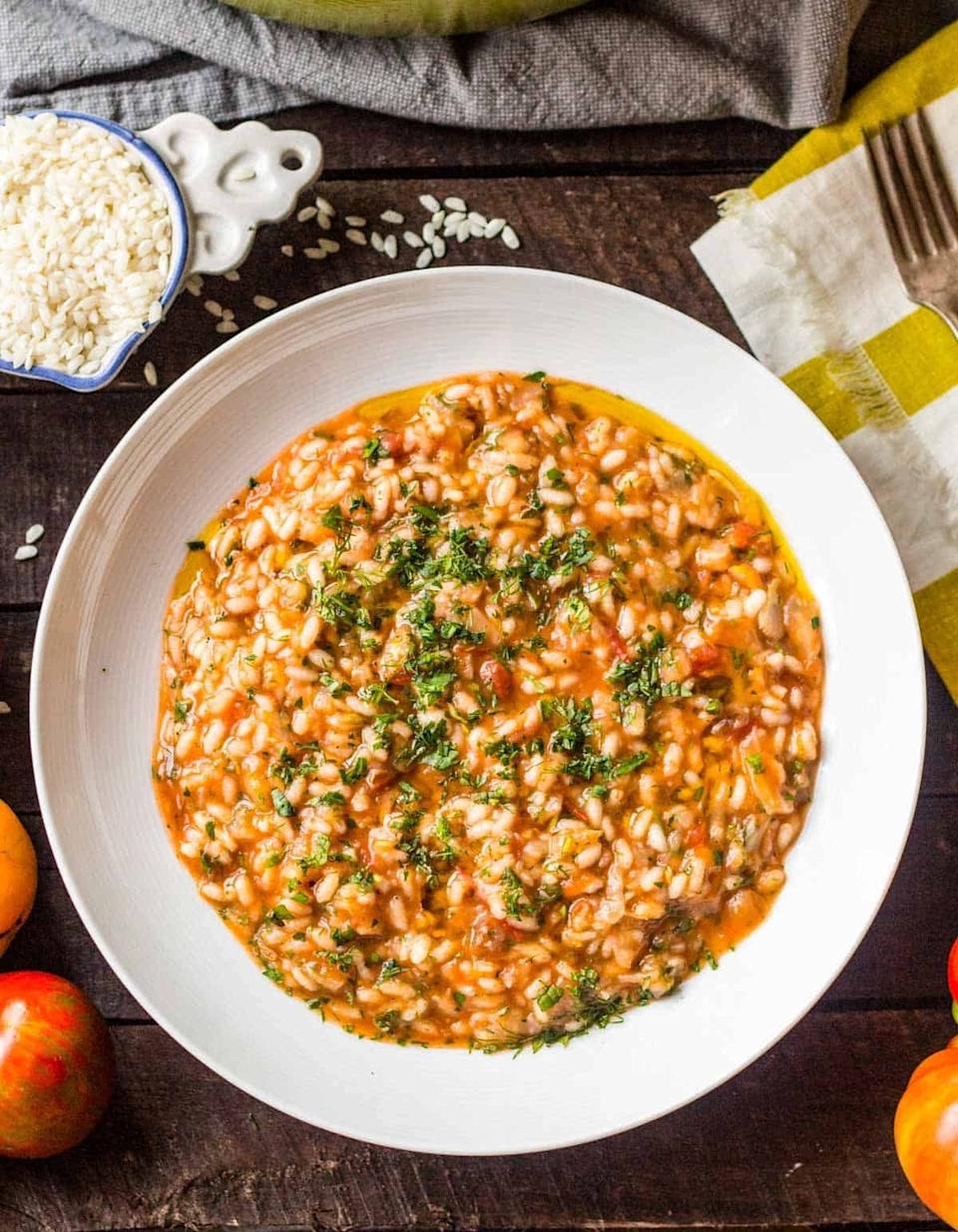 A plate of tomato risotto garnished with herbs, placed on a wooden table next to raw rice and apples