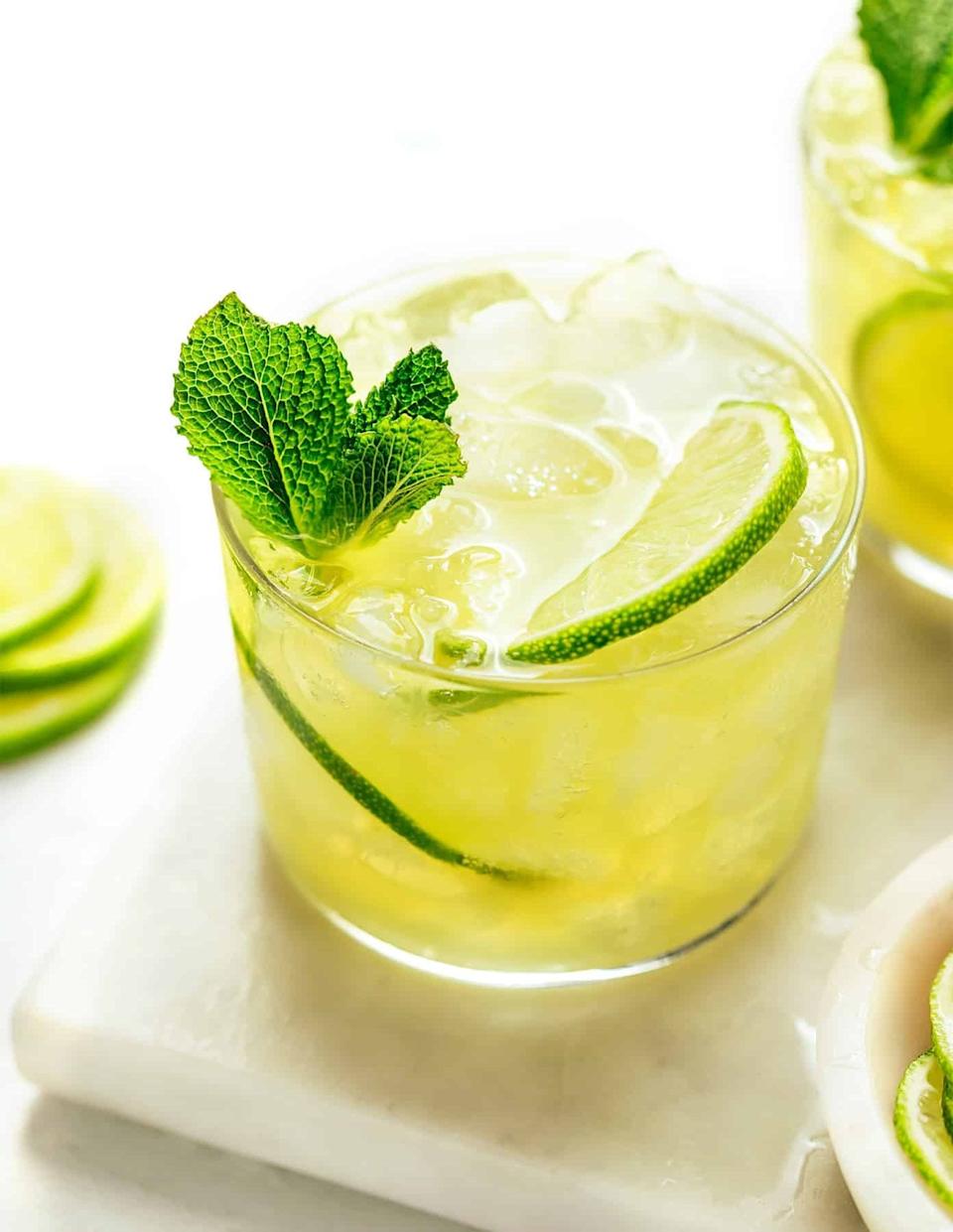 A refreshing iced cocktail garnished with mint and lime slices on a white surface, with additional lime slices nearby