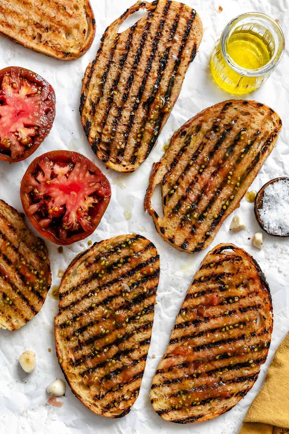 Grilled bread slices with olive oil, halved tomatoes, and garlic cloves on parchment paper