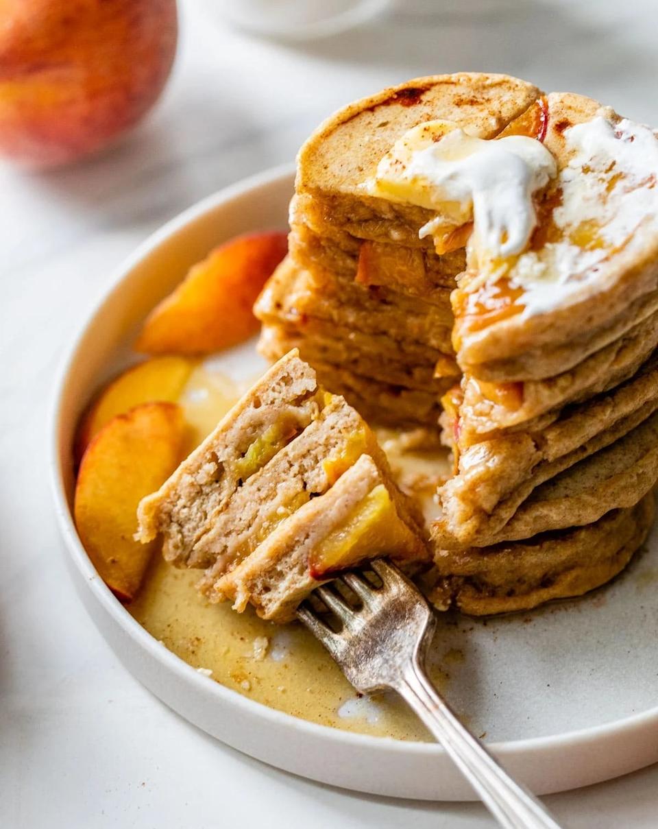 Stack of pancakes with fruit slices, butter, and syrup on a plate. A fork holds a cut portion, showing a close view of texture