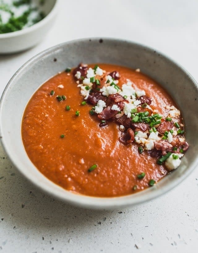 A bowl of creamy tomato soup garnished with crumbled cheese, chives, and diced onions on a speckled countertop