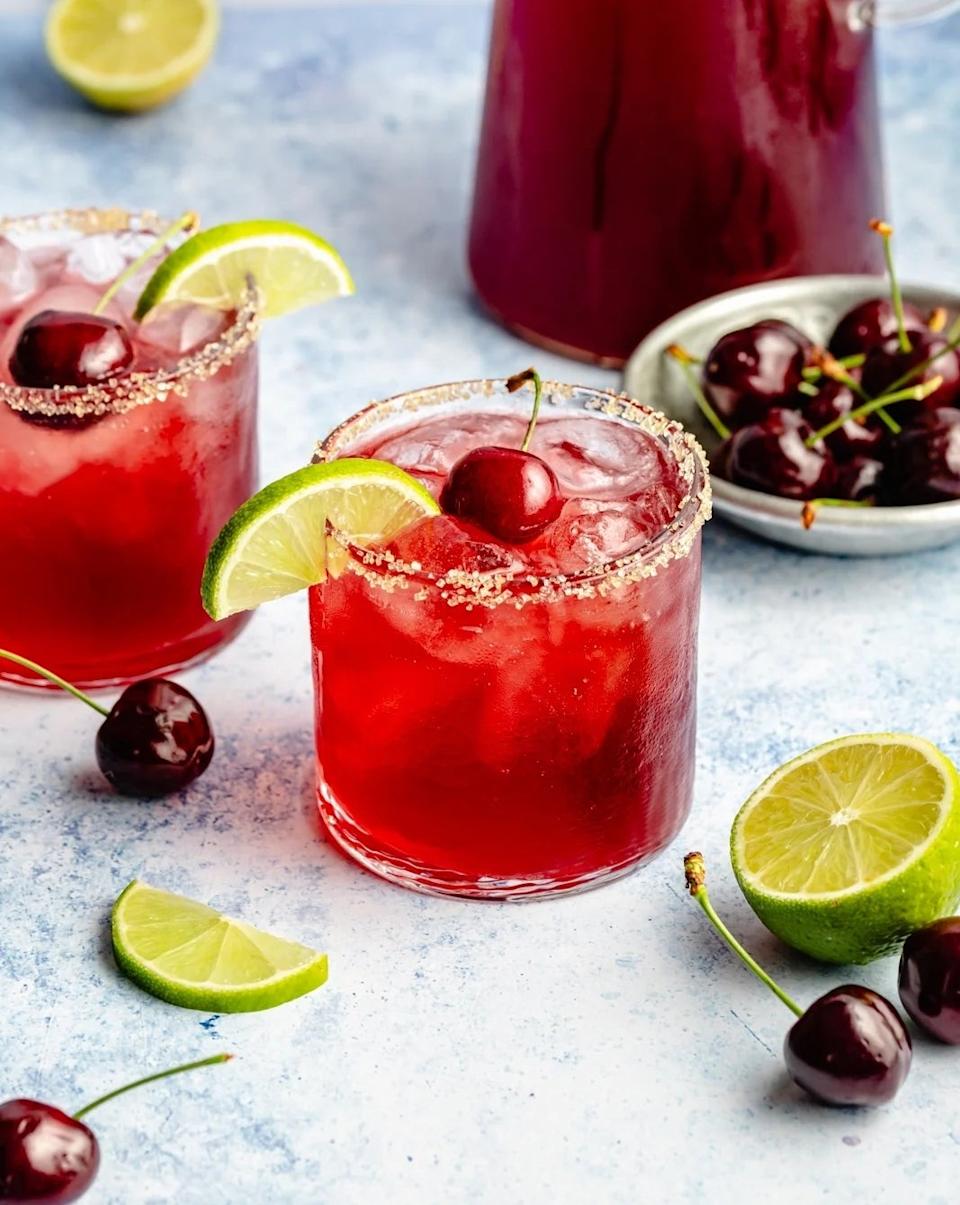 Two cherry cocktails with lime slices and sugared rims. Cherries and lime wedges are scattered around; a pitcher is in the background