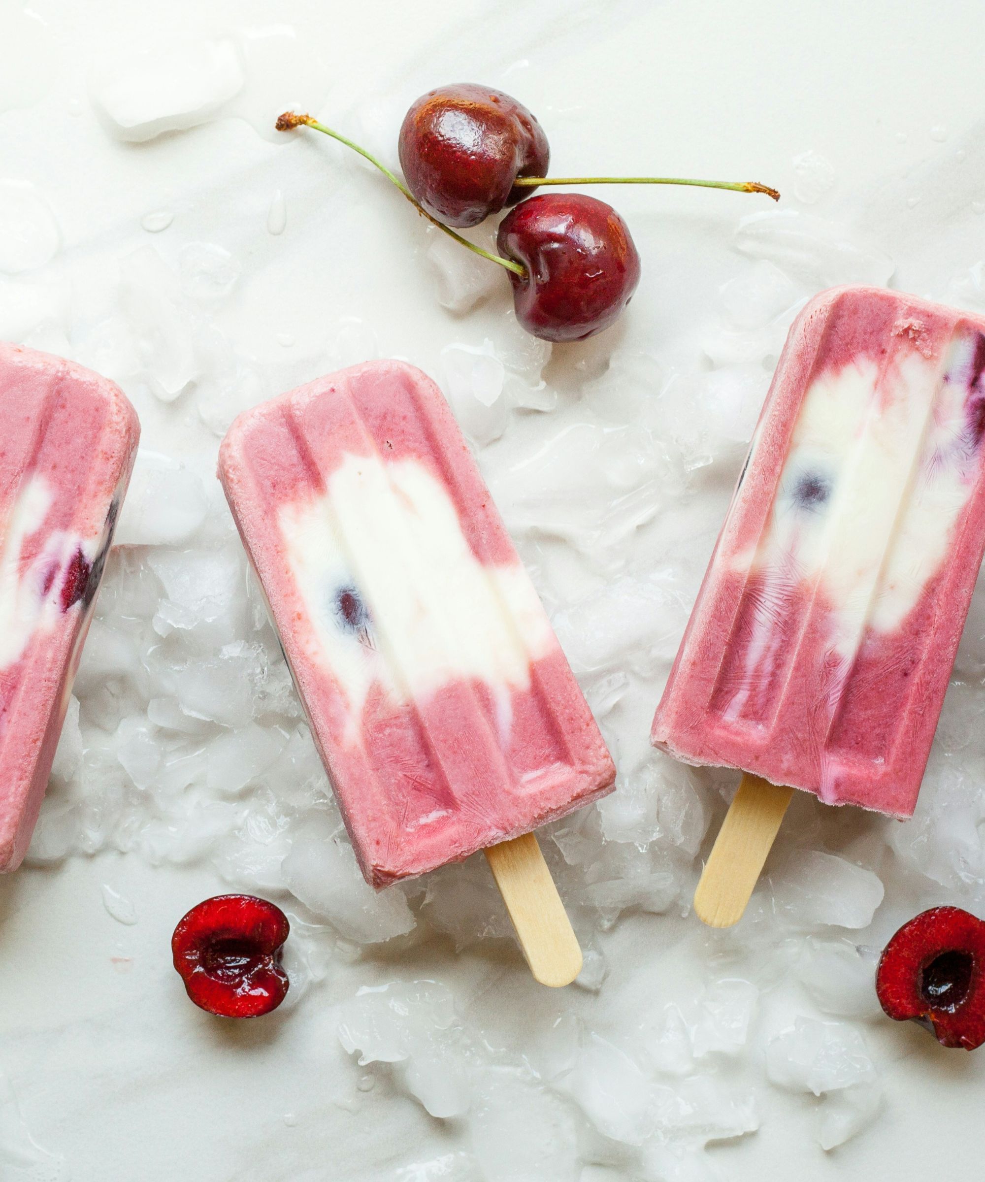 Cherry and vanilla popsicles on a white marble background, with pitted cherries scattered around them