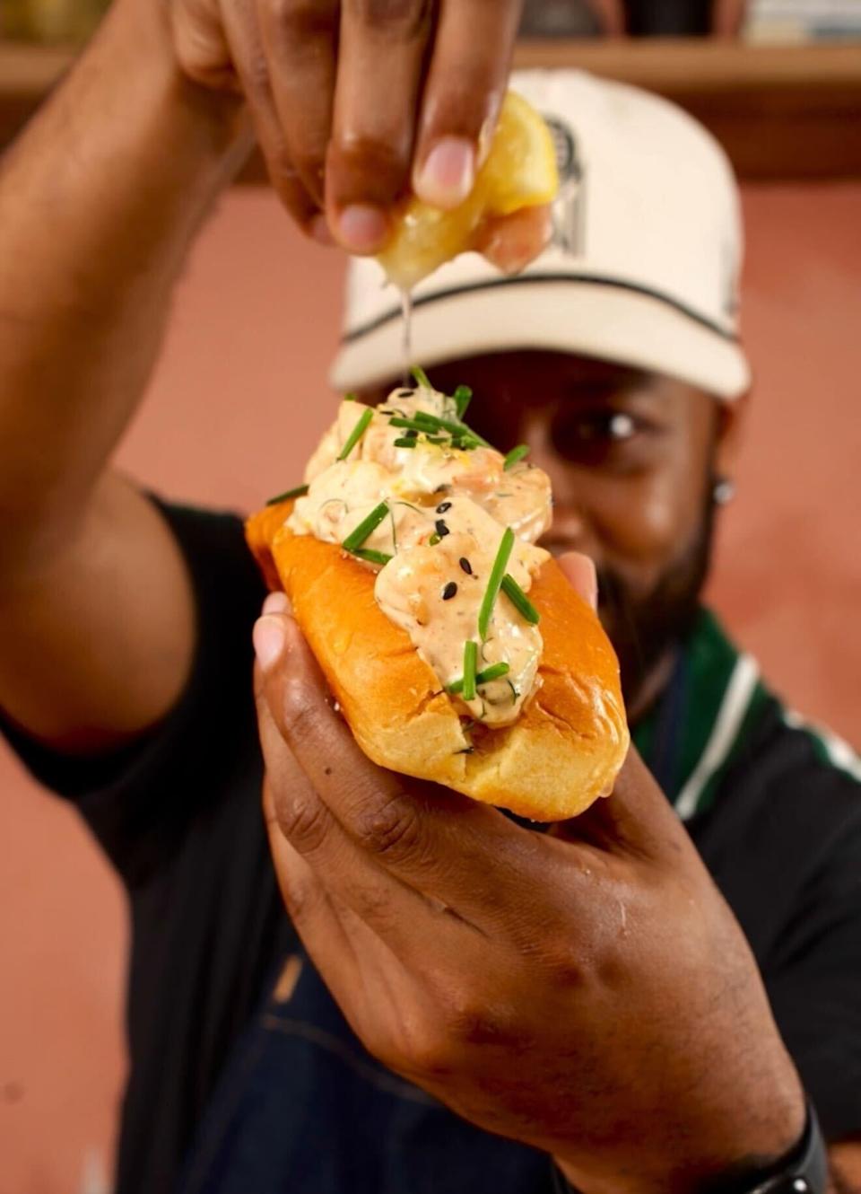 A person squeezes lemon over a gourmet lobster roll topped with chives and black sesame seeds, held up close to the camera