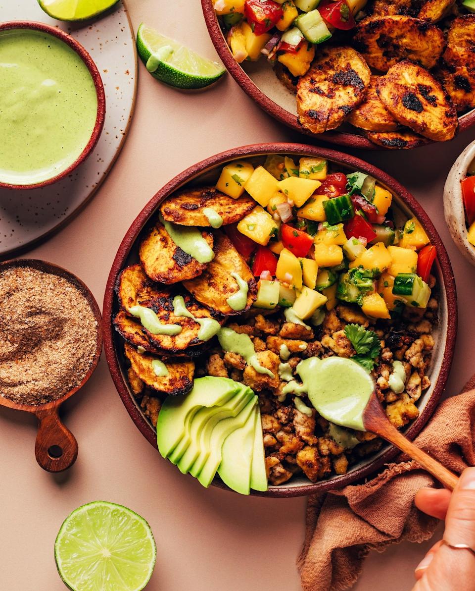 Bowl with grilled plantains, avocado slices, mango salsa, and a creamy sauce; side of lime wedges and a small bowl of dressing