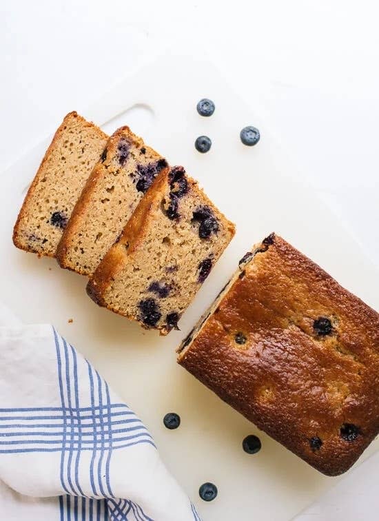 Blueberry loaf cake, partially sliced, on a white surface. Scattered blueberries and a striped cloth are nearby