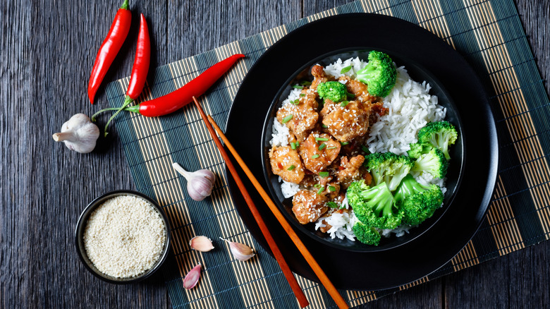Bowl of General Tso's chicken over rice with broccoli placed on a mat with chilies, garlic, and sesame seeds