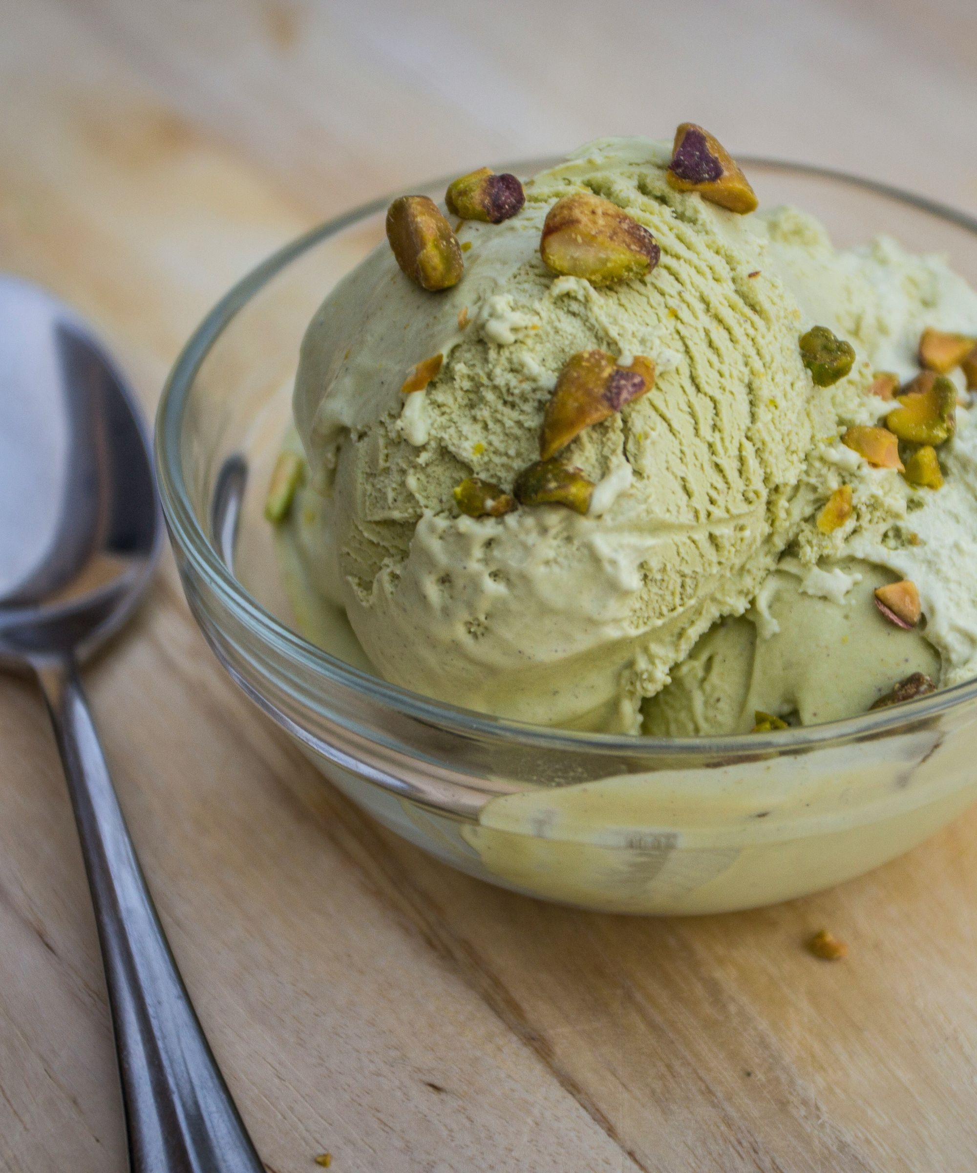 Glass bowl of pistachio ice cream with crushed pistachios on top, placed on a wooden table with a silver spoon beside it