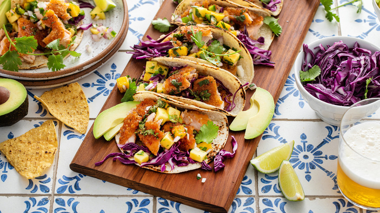 Fish tacos presented on wooden board with tortilla chips, beer, lime, cabbage and more on the side.