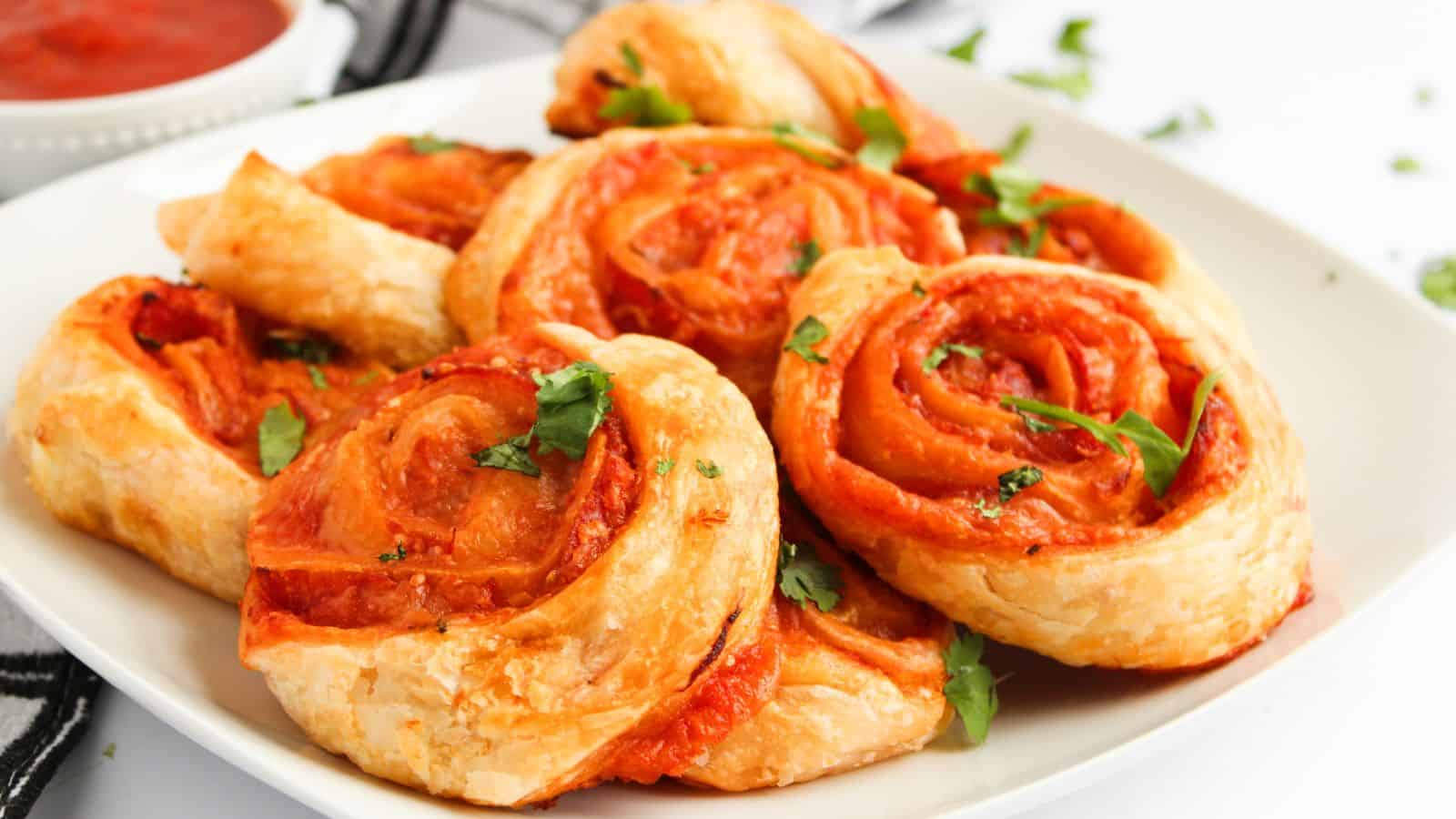 A plate of golden-brown pinwheel pastries filled with tomato sauce and garnished with fresh herbs. A small bowl of sauce is in the background.