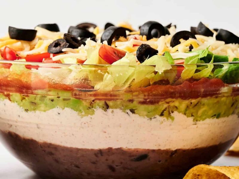A clear glass bowl showing layered dip with refried beans, sour cream, guacamole, lettuce, tomatoes, shredded cheese, and sliced black olives. Tortilla chips are visible in the foreground.