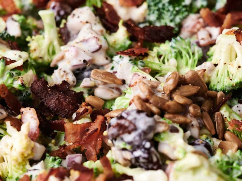 A close-up of a broccoli salad with creamy dressing, sunflower seeds, bacon pieces, and raisins.