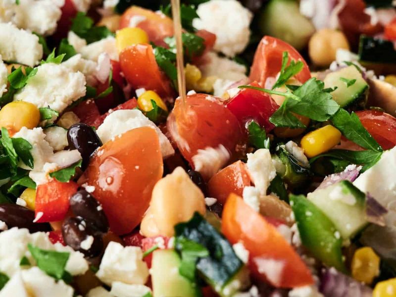 A close-up of a chopped salad with tomatoes, cucumbers, black beans, chickpeas, corn, feta cheese, parsley, and a drizzle of dressing.
