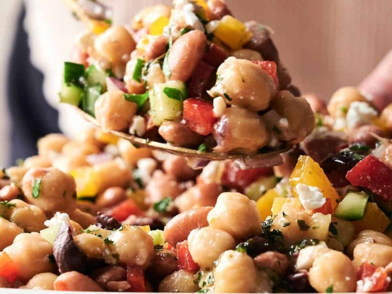 A close-up of a spoon scooping a chickpea salad with chopped cucumbers, tomatoes, bell peppers, beans, olives, and crumbled cheese.