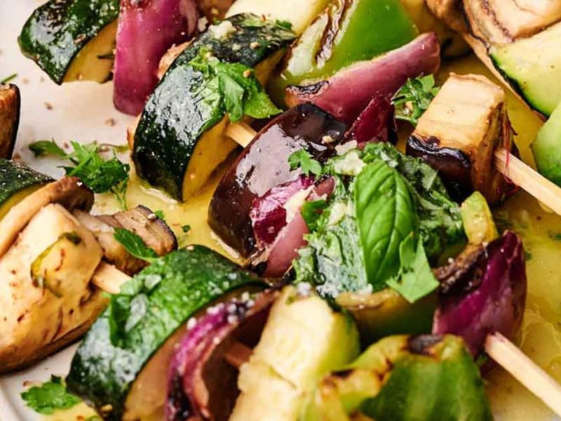 Close-up of vegetable skewers with zucchini, red onion, green bell pepper, and eggplant, garnished with fresh herbs.