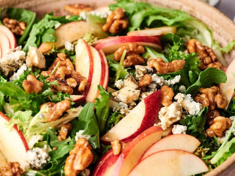 A salad with mixed greens, sliced red apples, walnuts, and crumbled blue cheese in a wooden bowl.