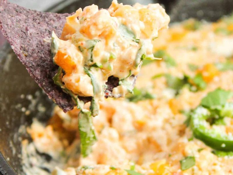 A close-up of a spoonful of cheesy dip with chopped green herbs and peppers, held above a bowl of the same mixture.