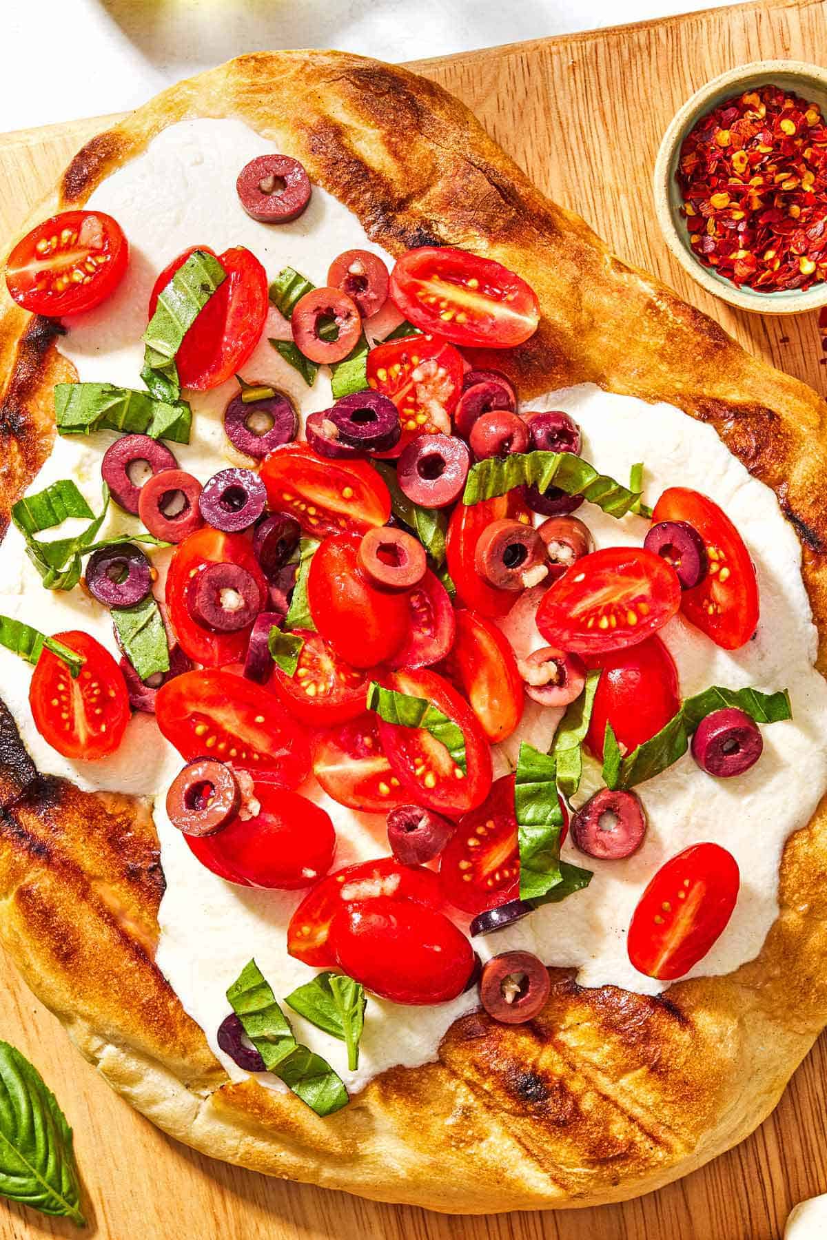 A grilled pizza on a wooden serving board with a bowl of red pepper flakes and some basil leaves.
