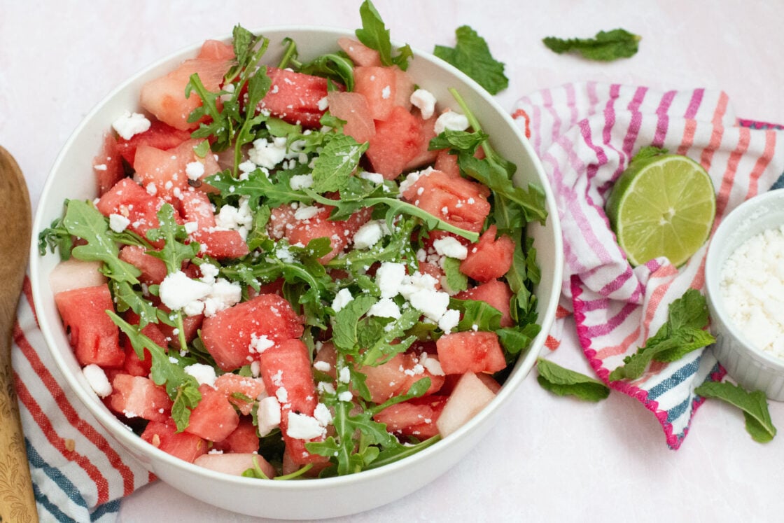 image: watermelon arugula and feta salad recipe with mint served in large white bowl on top of colorful striped towel, along side a wooden spoon, halved lime, and small white bowl with extra feta via Organic Authority