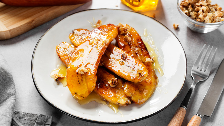 Sauteed Bananas Foster with ice cream, honey and walnuts served on a white plate with utensils on the side.