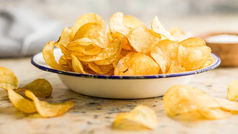 Potato chips in a white bowl with a blue rim with some spilled out on the table