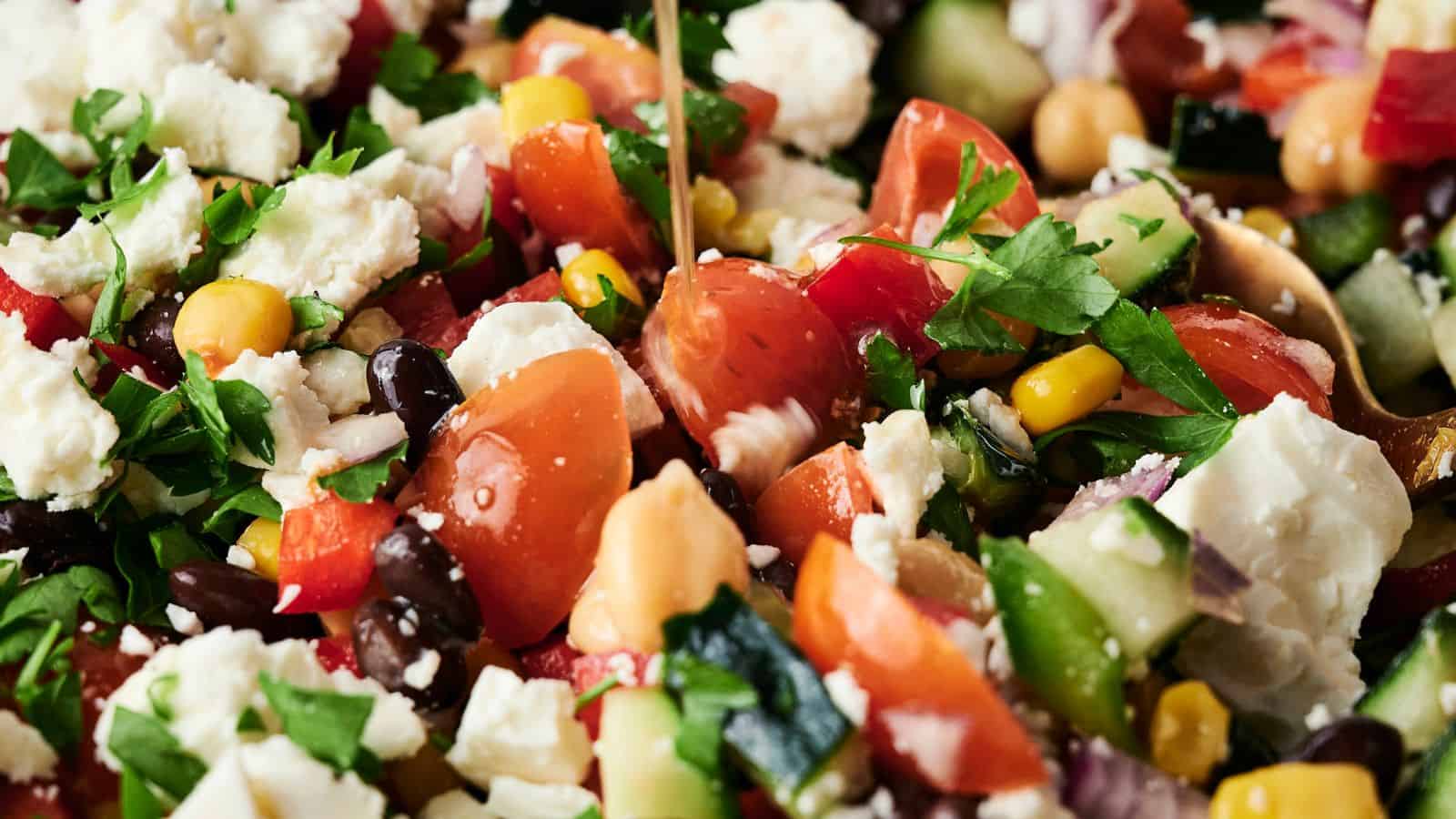 Close-up of a colorful salad with cherry tomatoes, cucumbers, black beans, chickpeas, corn, red onion, parsley, and crumbled feta cheese, being drizzled with dressing.