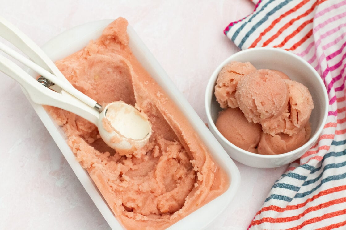 Image: Pink watermelon sorbet or ice cream scooped into a white bowl with white ice cream scoop out of a white enamel bread pan with colorful striped towel off to right side via Organic Authority.