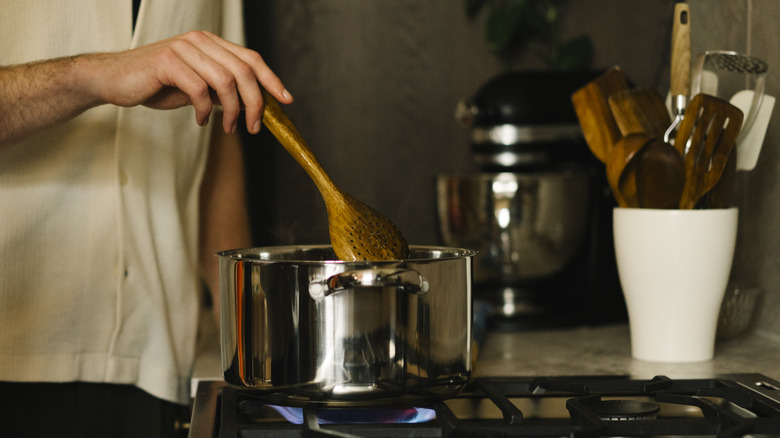 A man holding a wooden spatula, stirring a stainless steel pot on a lit stove